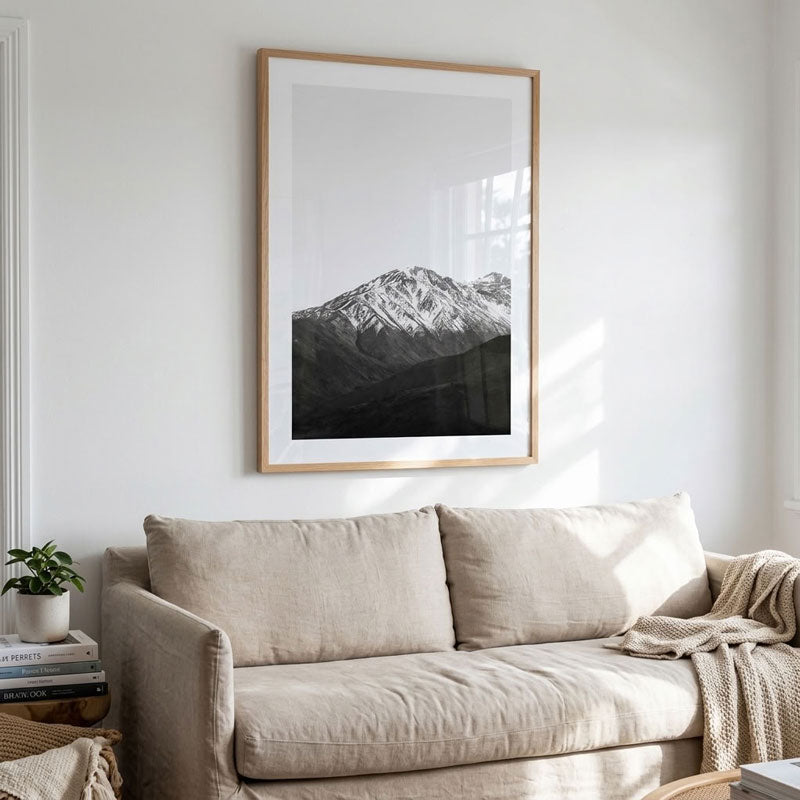 A large framed black and white mountain landscape print hangs on a white wall above a beige sofa with a knit throw blanket.