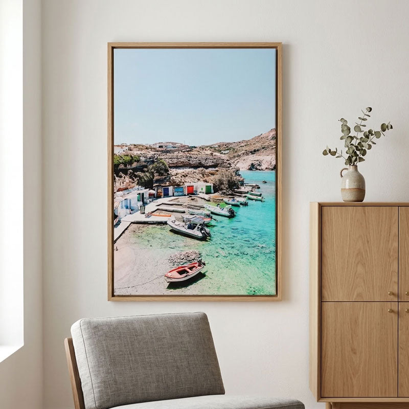A framed vertical art print depicting a scenic coastal harbor with turquoise water, boats, and rocky hills, displayed on a light beige wall above a gray chair and next to a wooden cabinet.