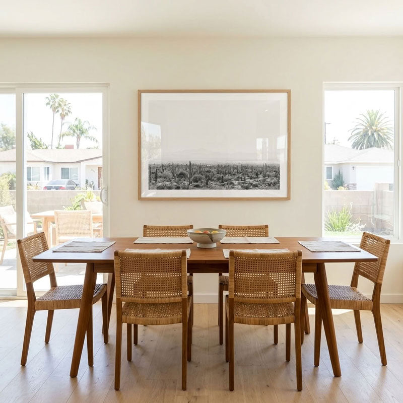 A dining room scene with a rectangular wooden dining table and six woven-back chairs, set in a bright room with large windows and a framed black-and-white landscape print on the wall.