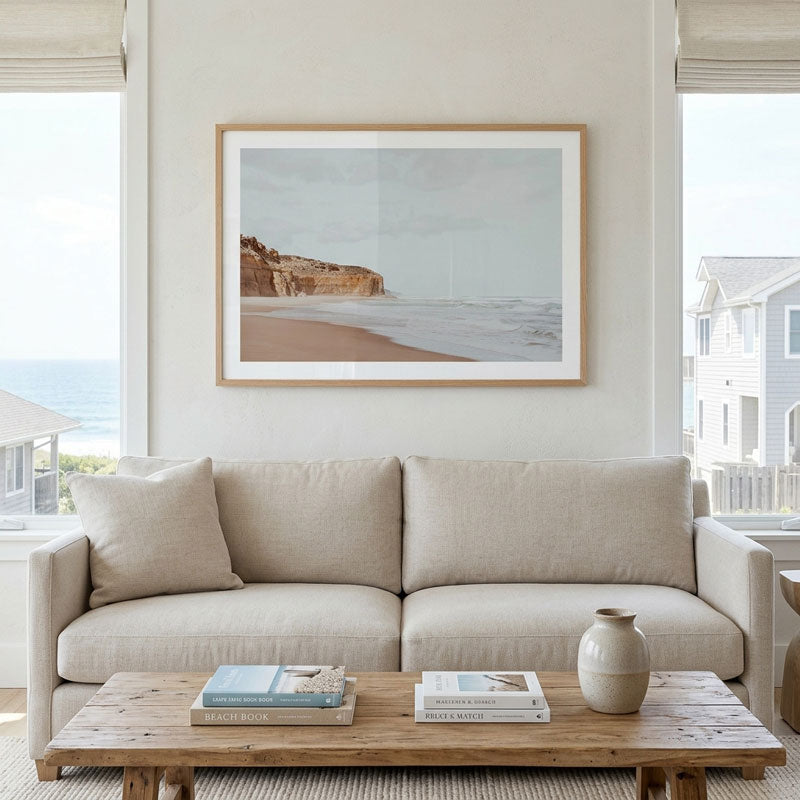A framed coastal landscape print hangs on a light beige wall above a cream sofa in a bright, modern living room. The print shows a beach and rocky cliff under a pale sky, with a light wood frame and white mat.