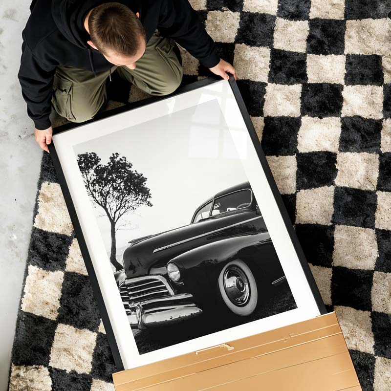 Person holding a framed black and white photograph of a classic car on a checkered floor.