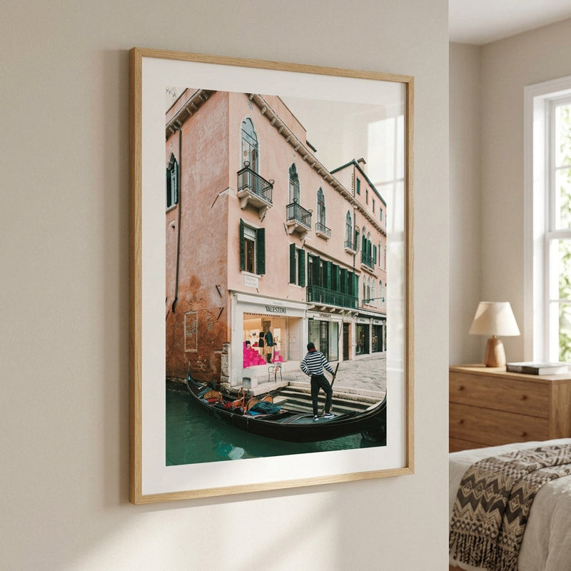A framed art print depicting a Venetian canal scene with a gondola and historic buildings, styled on a beige wall in a modern bedroom setting.