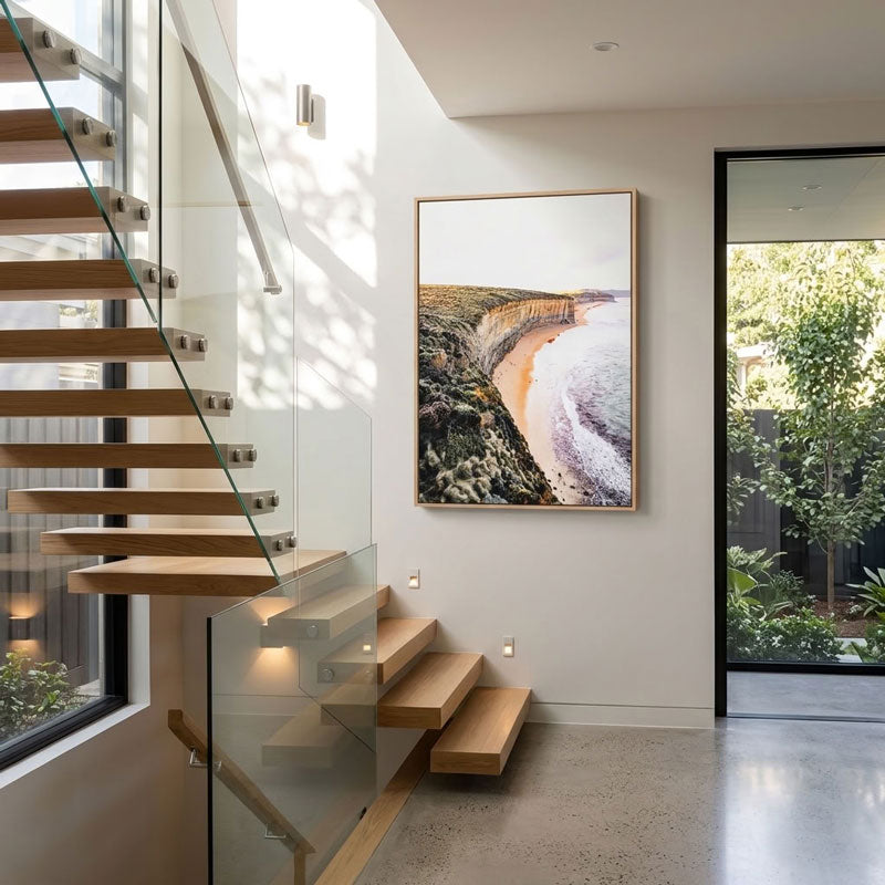 A framed vertical landscape photograph of a coastal cliff and beach print hangs on a white wall in a modern interior with floating wooden stairs and glass railings.