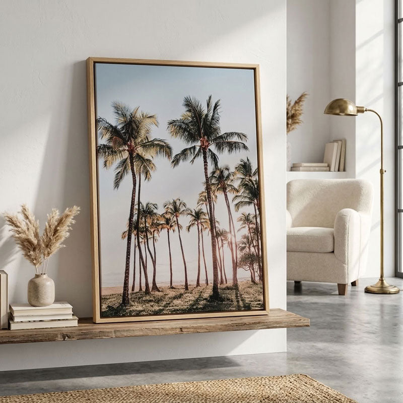 A framed vertical art print featuring a tropical beach scene with palm trees, styled in a modern living room setting on a wooden shelf next to a white armchair and a brass floor lamp.