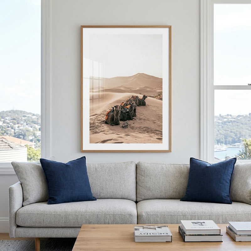 A framed vertical art print depicting a desert landscape with camels, hung on a white wall above a modern light gray sofa with navy blue pillows in a bright living room.