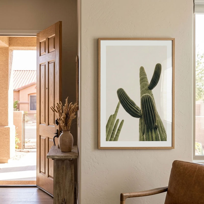 A framed botanical art print of a green saguaro cactus on a white background, styled on a beige wall in a modern home entryway next to an open wooden door and a leather chair.
