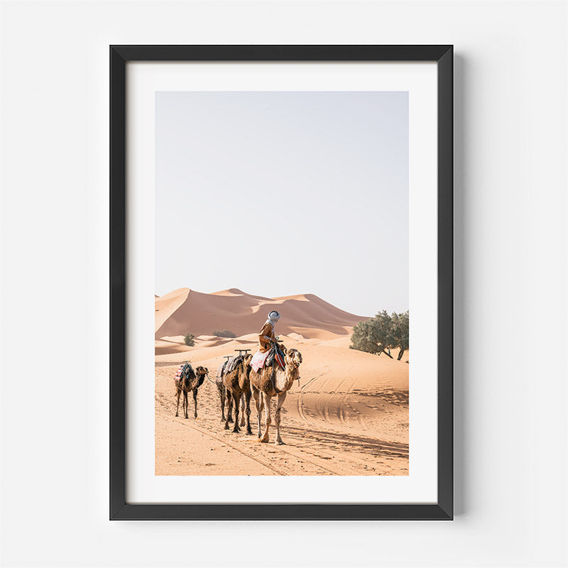 Framed photograph of a person riding a camel in the desert with sand dunes in the background.
