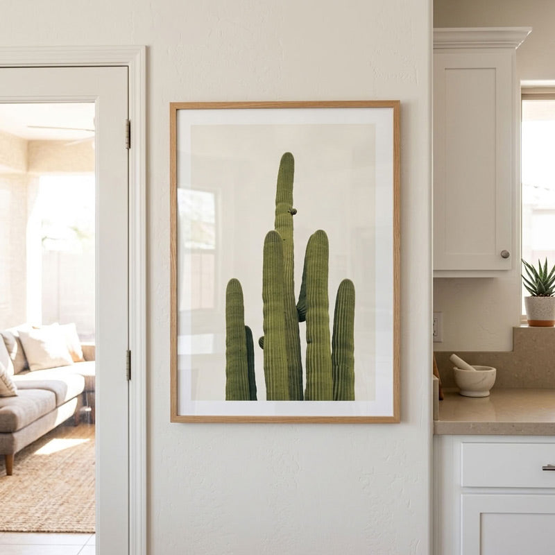 A framed art print of a green saguaro cactus on a white background, hanging on a beige wall in a bright, modern kitchen.