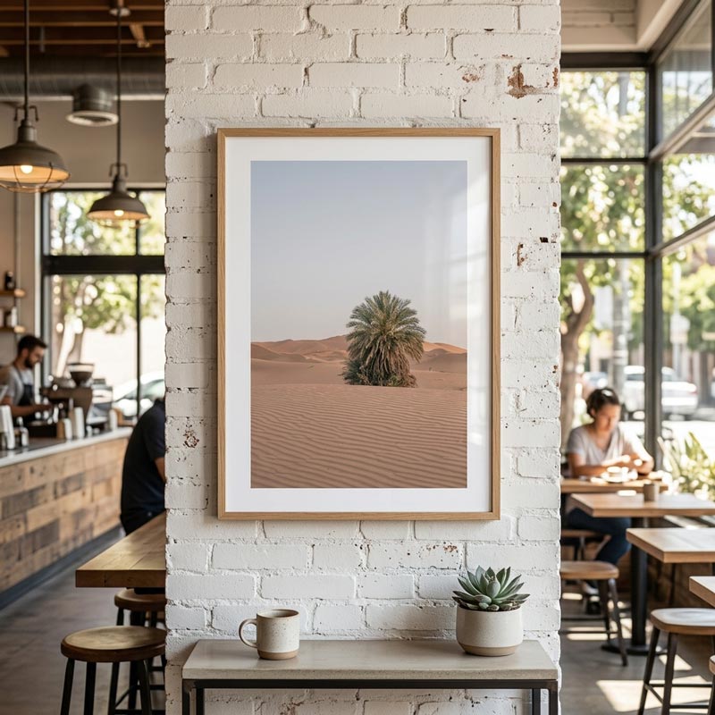 A framed desert landscape art print featuring a single palm tree on sand dunes, displayed on a white brick wall in a modern cafe interior.