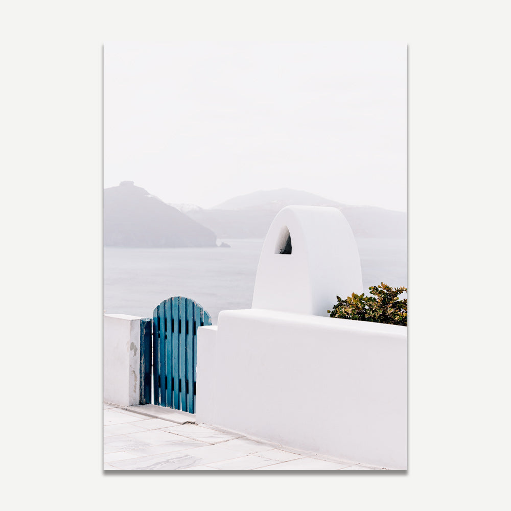 A wall art print featuring a white washed gate and architecture in Santorini with a blue gate, leading to a view of the ocean in the background.