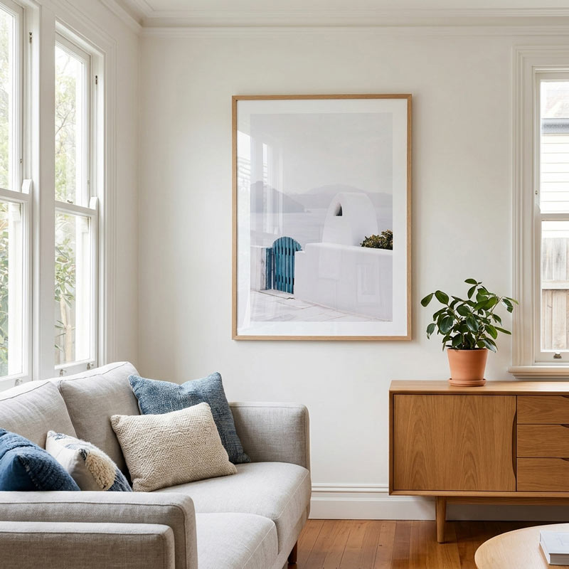 A framed minimalist art print depicting white buildings and a blue draped textile, hanging on a cream wall in a bright, modern living room with a light gray sofa and a wooden sideboard.