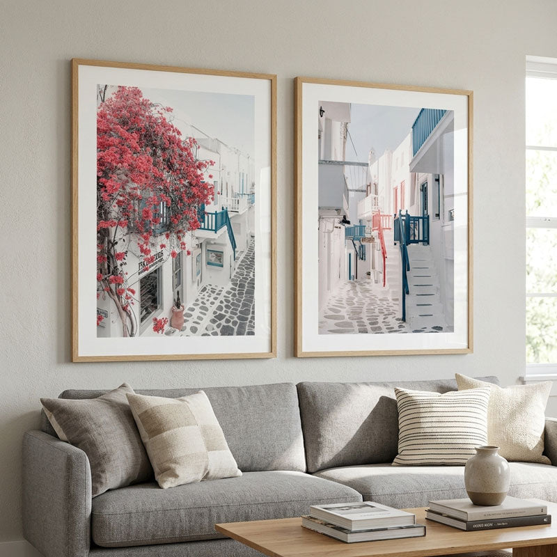 Two framed art prints depicting Mediterranean street scenes with white buildings, blue accents, and red flowers, displayed on a light gray wall above a gray sofa with neutral throw pillows.