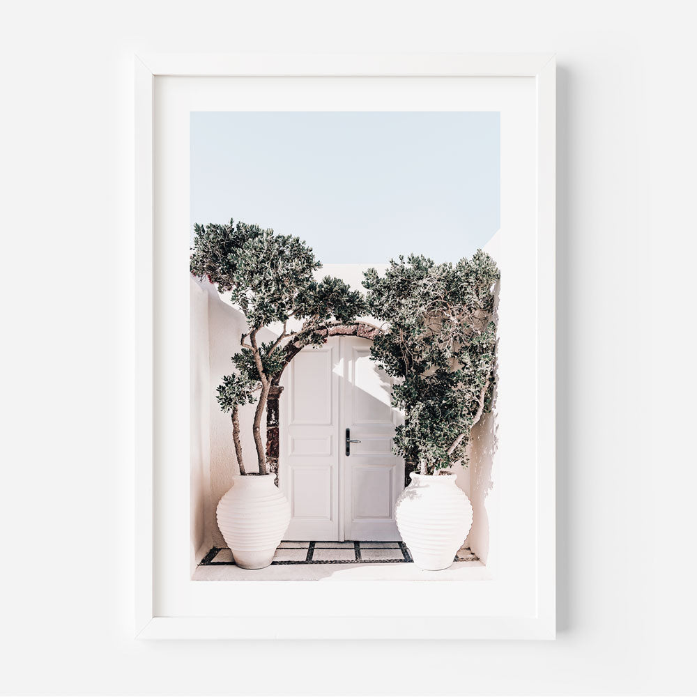 A framed photograph of two white pots of plants placed in front of a white door, with greenery peeking from behind, captured in a coastal setting.