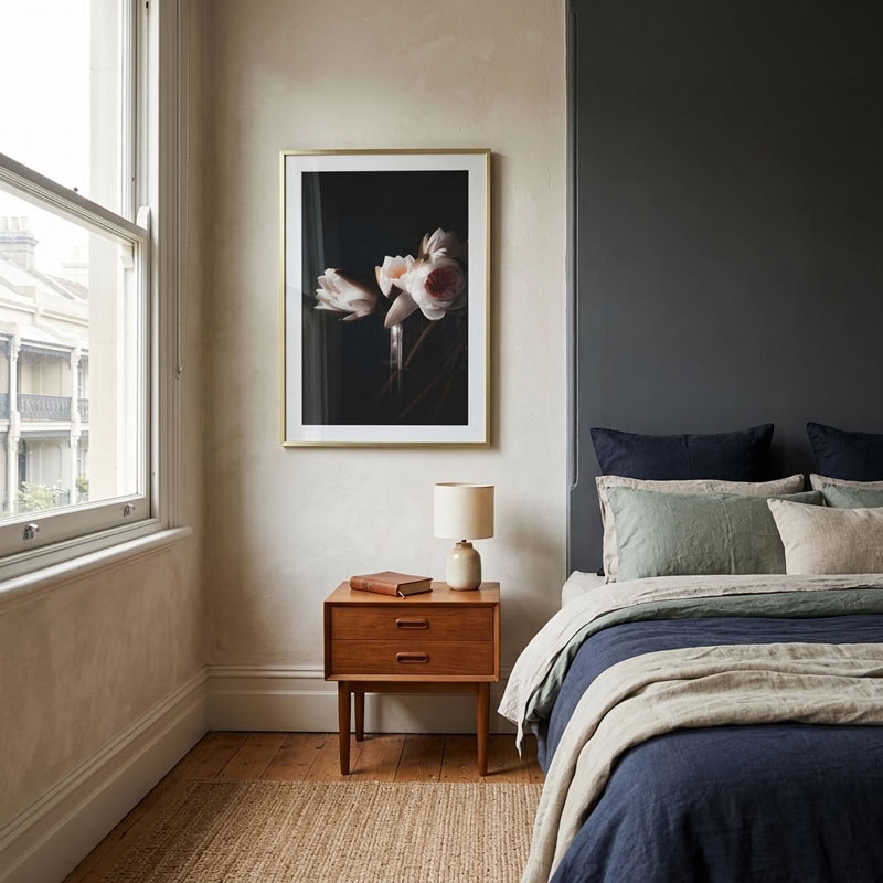A framed black and white floral art print hangs on a beige wall in a styled bedroom next to a wooden nightstand and a bed with blue and green bedding.