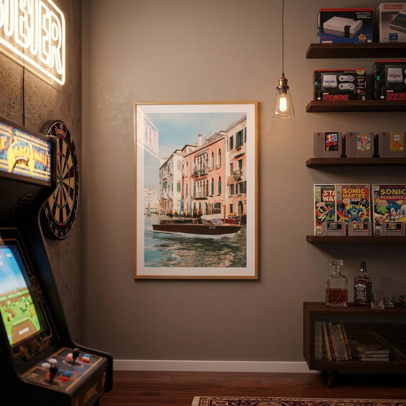 A framed art print depicting a Venice canal scene with colorful buildings and a boat, displayed on a gray wall in a retro-themed room with arcade machines, shelves, and a hanging light.