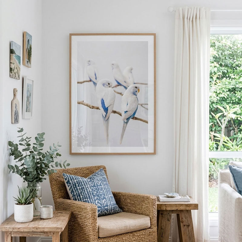 A framed wall art print featuring four white birds with blue accents perched on branches, displayed in a bright living room above a wicker armchair with a blue patterned pillow.
