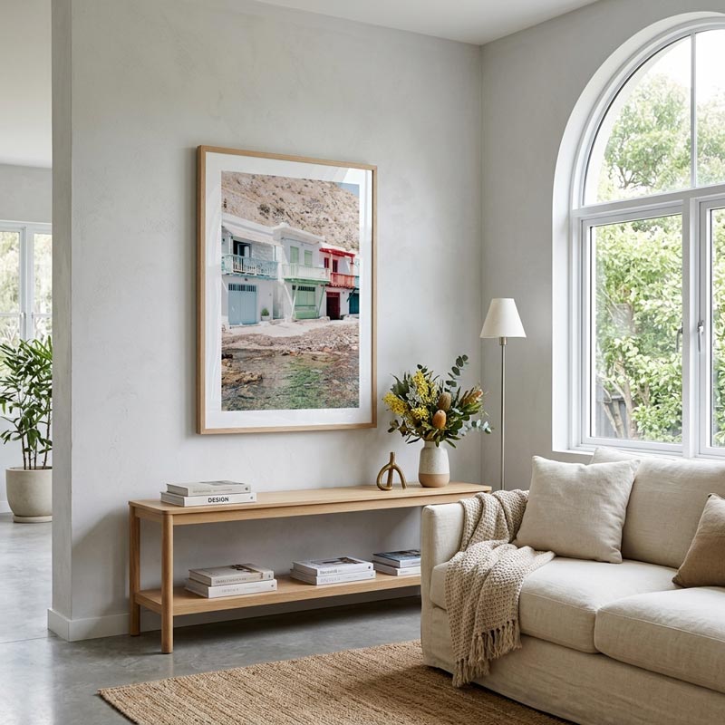 A framed art print depicting coastal houses by the water, styled on a light wood console table in a bright, modern living room with a beige sofa and large arched window.