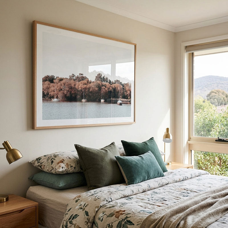 A framed landscape art print of a lake with trees and boats hangs on a beige wall above a bed with green and floral pillows in a modern bedroom setting.