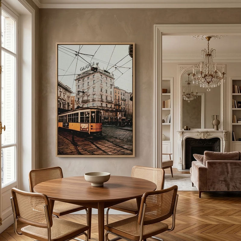 A framed vertical art print depicting a vintage yellow tram on a cobblestone street in front of a building with a 'G. Calmette' sign, displayed on a beige wall in a beautifully styled dining room with a wooden table, woven chairs, and a chandelier visible in the background.