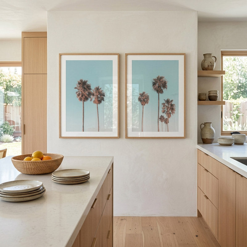 Modern kitchen with light wood cabinets, white countertops, and framed palm tree prints on the wall.