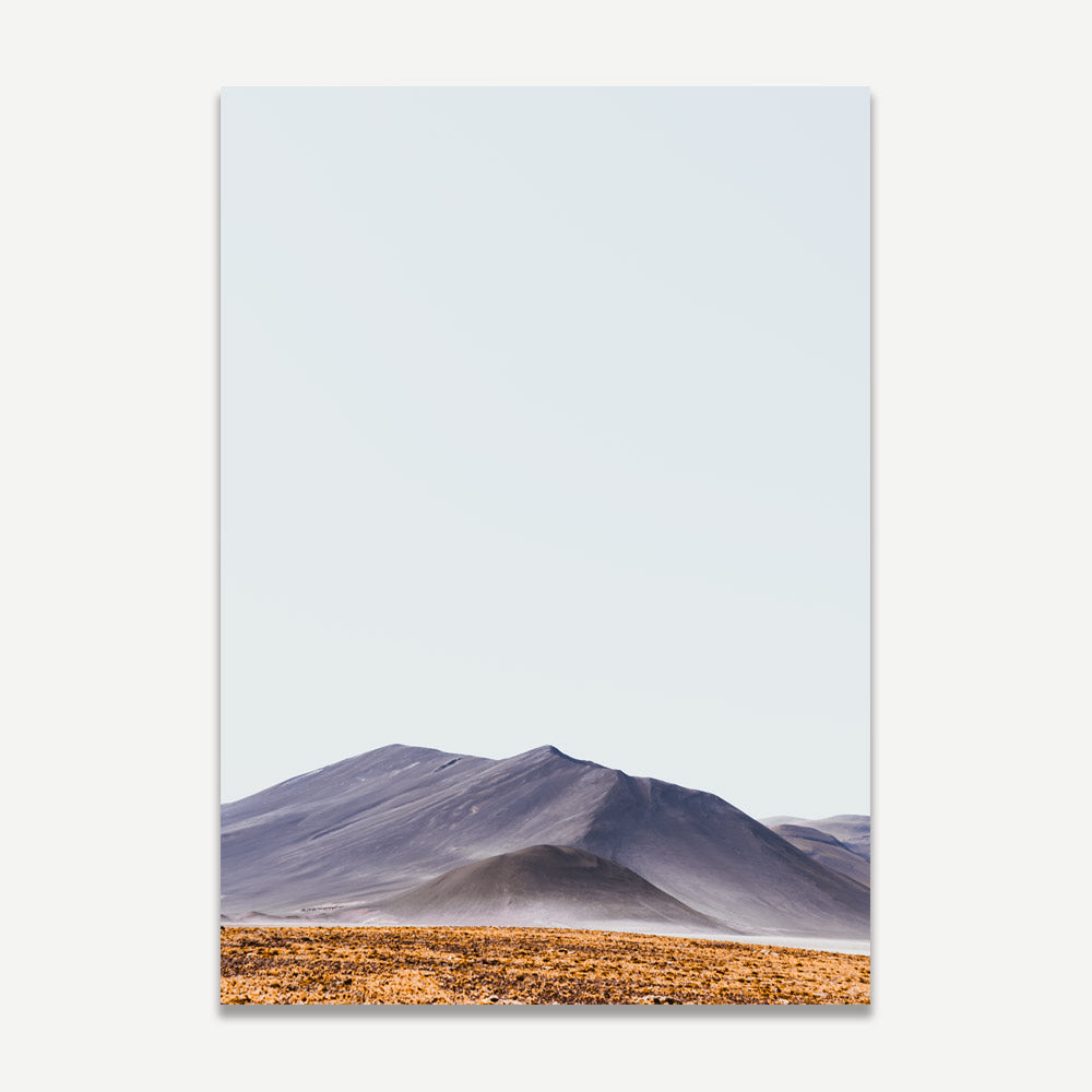 A landscape photograph featuring a clear blue sky, with orange and gray tones over a desert plain leading up to a range of mountains.