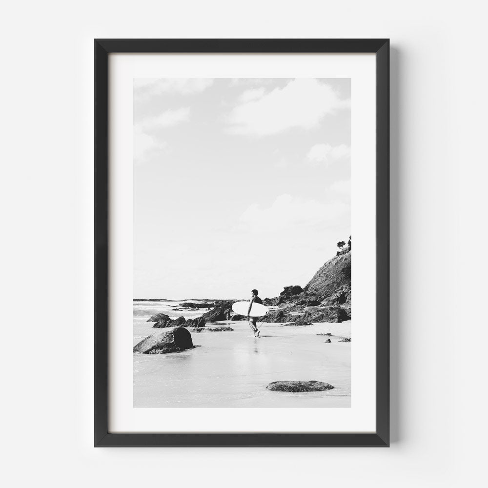 Black and white photograph of a person surfing, with rocks and a cliff in the background, framed in a black frame.