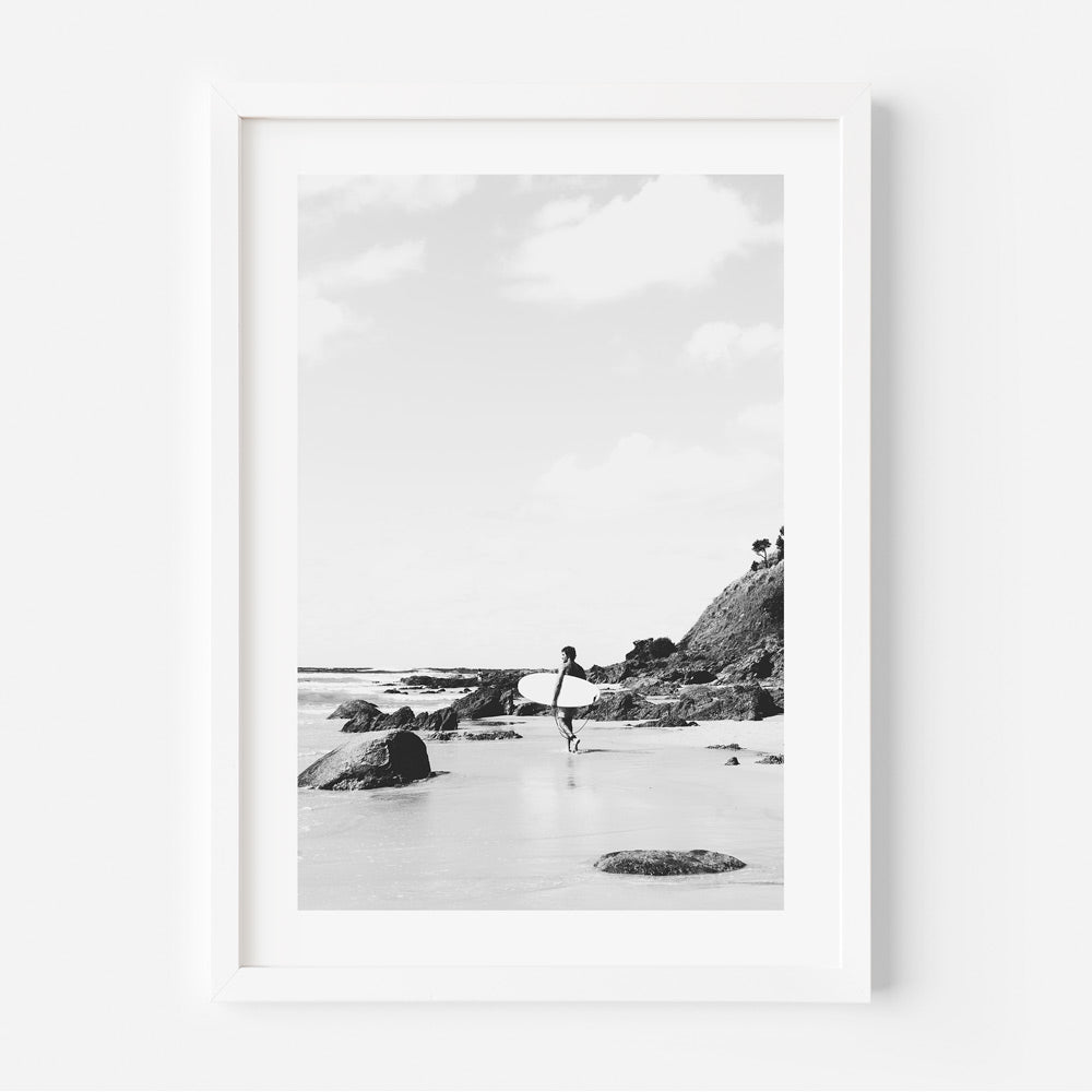 Black and white photograph of a person surfing, with rocks and a cliff in the background, framed in a white frame.