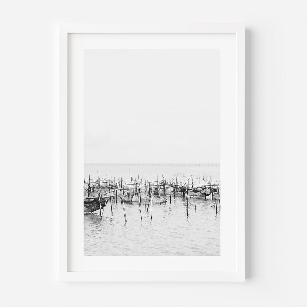A black and white photograph of a fishing village with fishing nets and wooden posts, captured in a framed image.