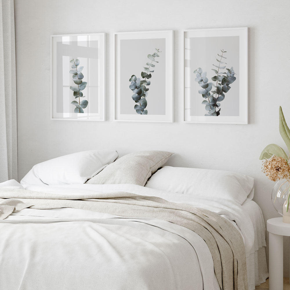 A set of three framed artwork pieces depicting gum leaves, hung on a white wall above a bed with white linens.