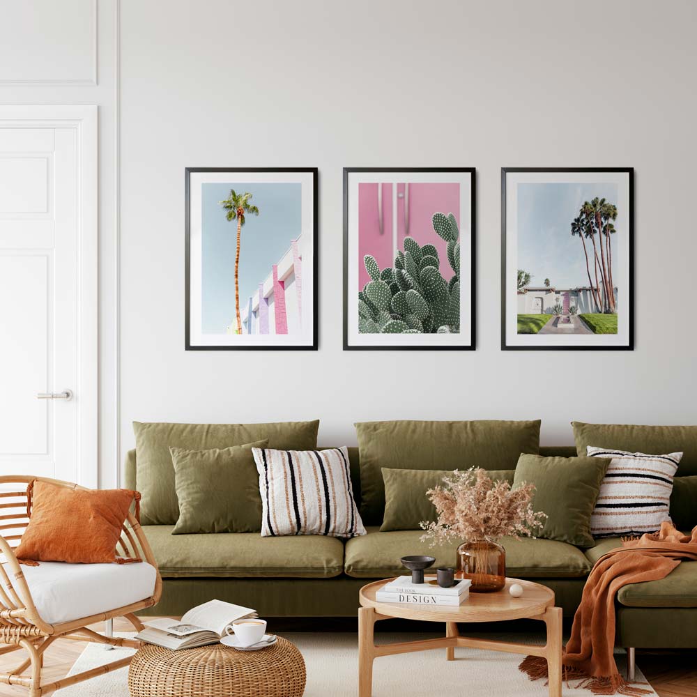 A set of three framed artworks hanging on a wall over a green sofa, featuring a palm tree, a pink cactus, and a pink door with palm trees in the background.