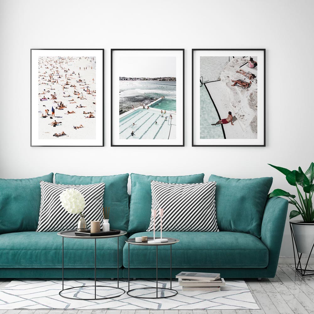 A set of three framed photographs with beach and poolside scenes, displayed above a green sofa with decorative pillows in a room with light walls and a plant.