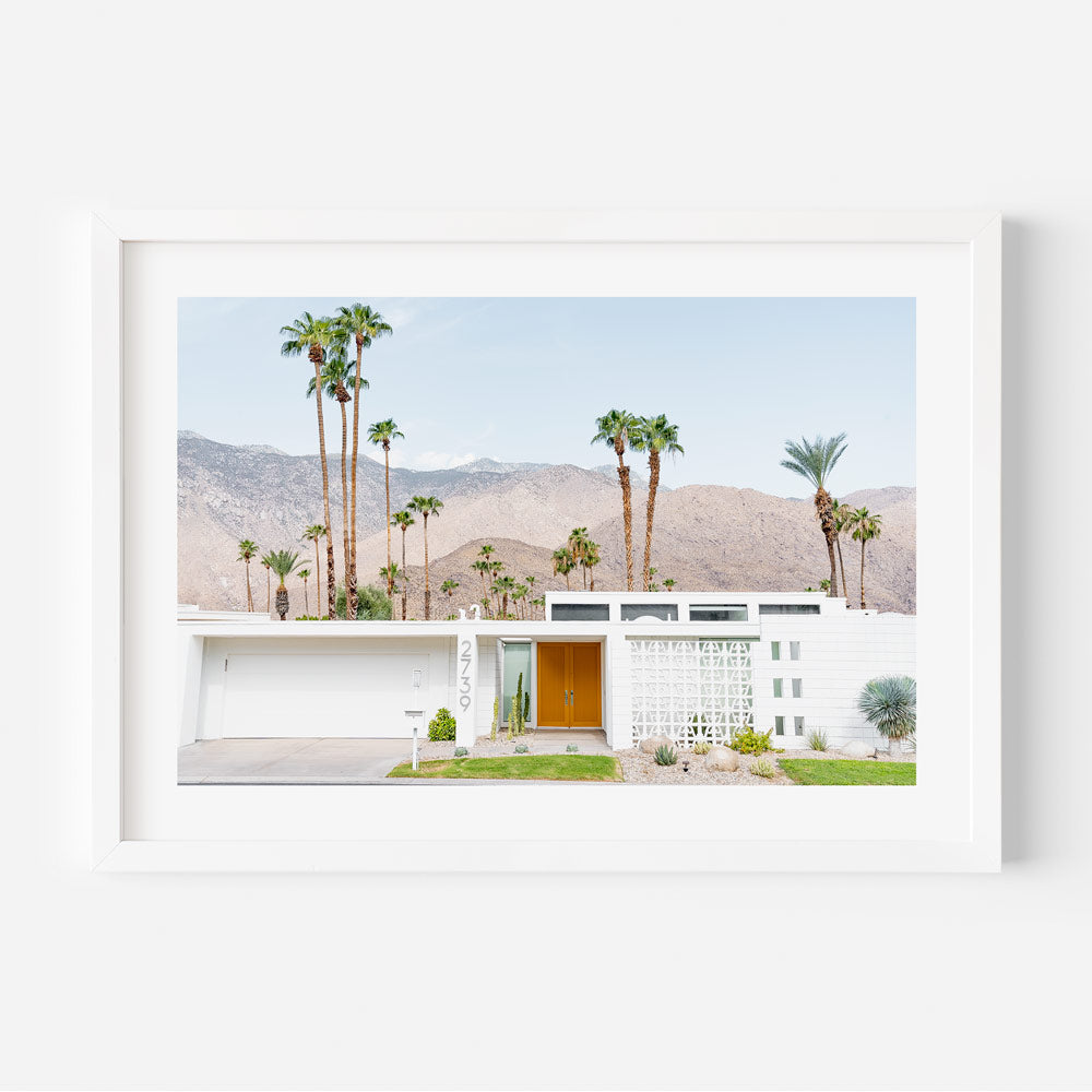 A photograph of a house with a striped orange and yellow door, set against a backdrop of palm trees and a mountain range.