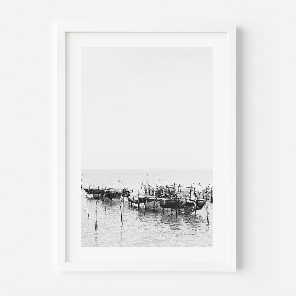 Black and white photograph of a fishing village with boats and wooden structures, captured within a white frame.