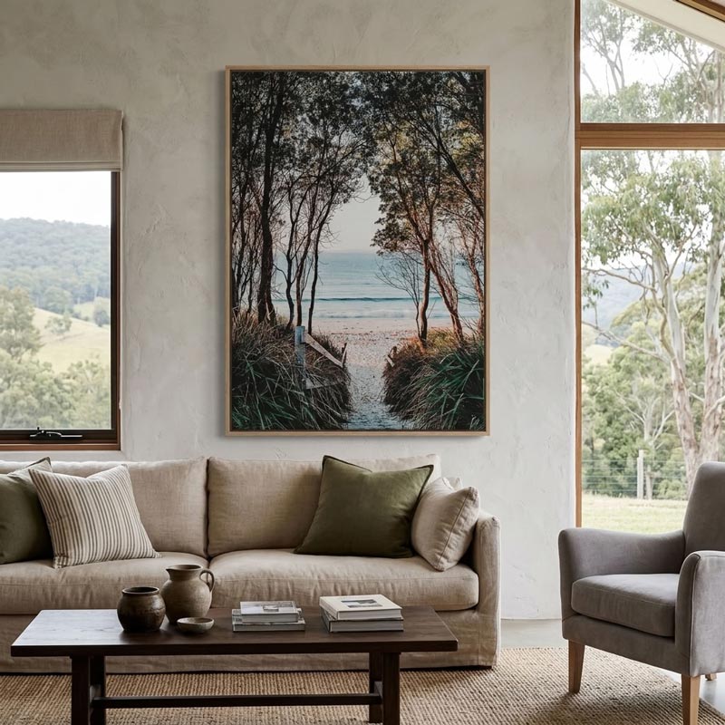 A framed vertical landscape art print depicting a forest path leading to the ocean, displayed on a textured white wall above a beige sofa in a modern living room setting.