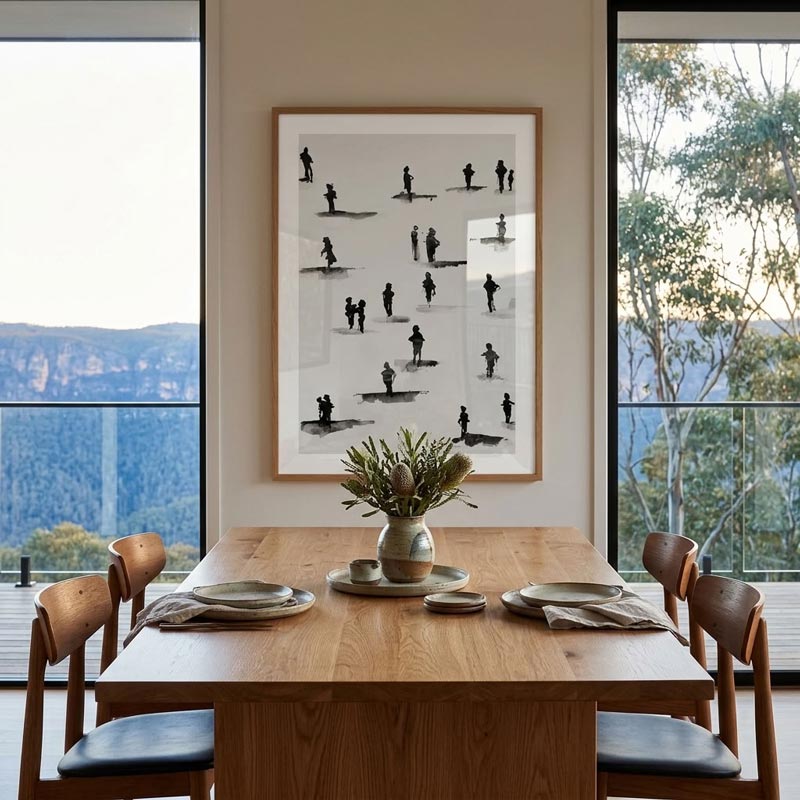 A framed black and white art print featuring minimalist human silhouettes, displayed on a beige wall above a wooden dining table in a modern dining room with large windows overlooking a scenic blue mountain landscape.