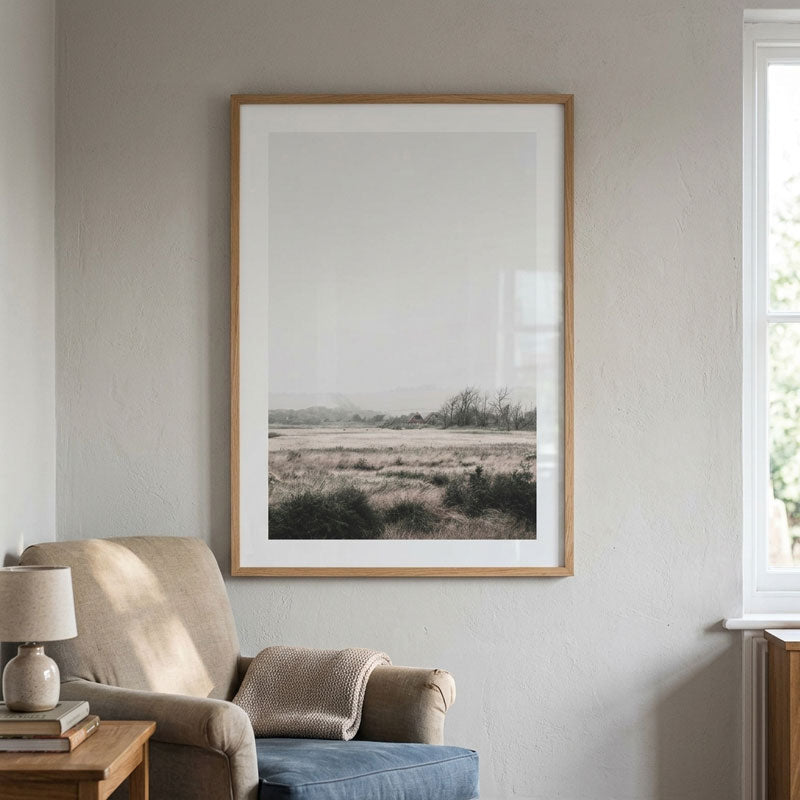A framed landscape art print with a light wood frame and white mat, hanging on a textured light gray wall above a beige armchair in a styled room.