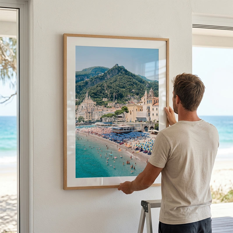 Man hanging a framed photograph of a coastal scene on a wall with ocean view.