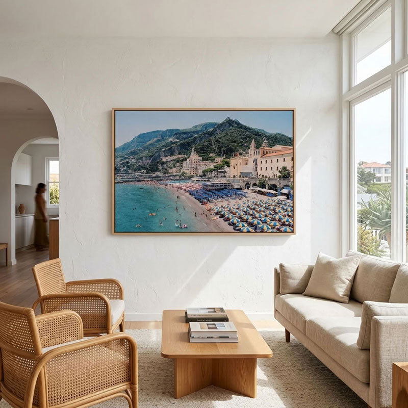 A large framed canvas print of a coastal town with a beach, blue water, and mountains, displayed on a white wall in a modern, sunlit living room with a sofa, chairs, and a coffee table.