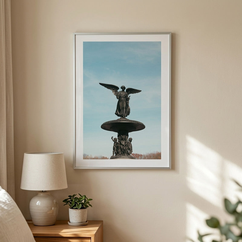 A framed art print of a winged angel statue on a pedestal against a blue sky, displayed on a beige wall in a styled bedroom setting next to a lamp and a small plant.