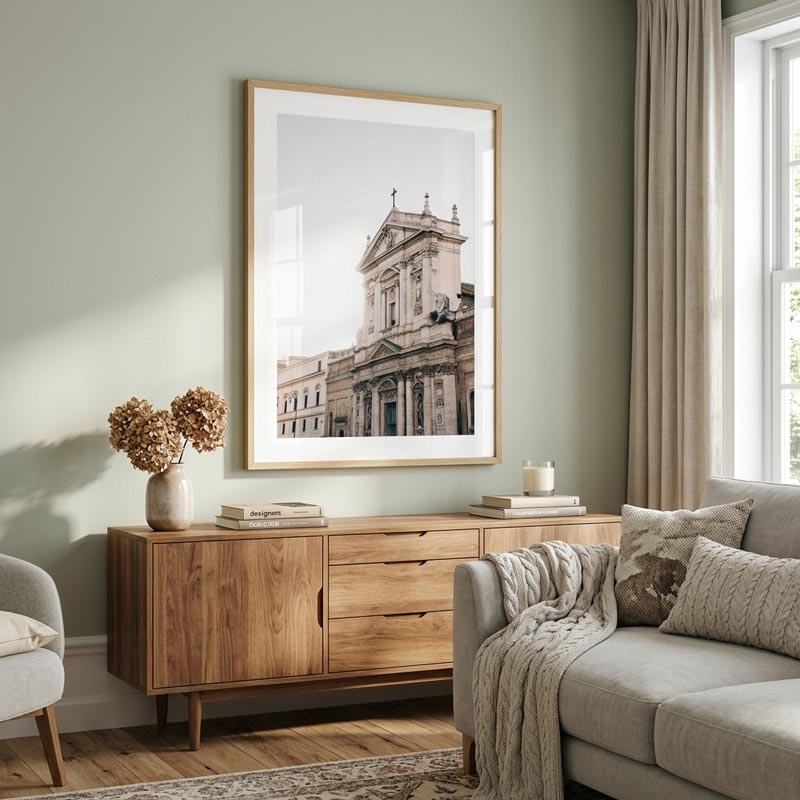 A large framed black and white architectural art print of a classical building, displayed on a sage green wall above a wooden sideboard in a styled living room setting.
