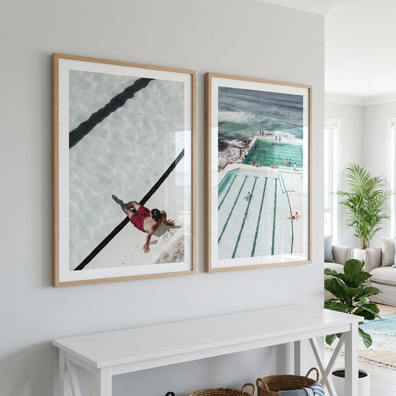 Two framed vertical art prints hanging side-by-side on a white wall above a white console table in a bright, modern room. The left print features an overhead shot of a swimmer in a pool, and the right print shows an aerial view of a swimming pool and beachside water.