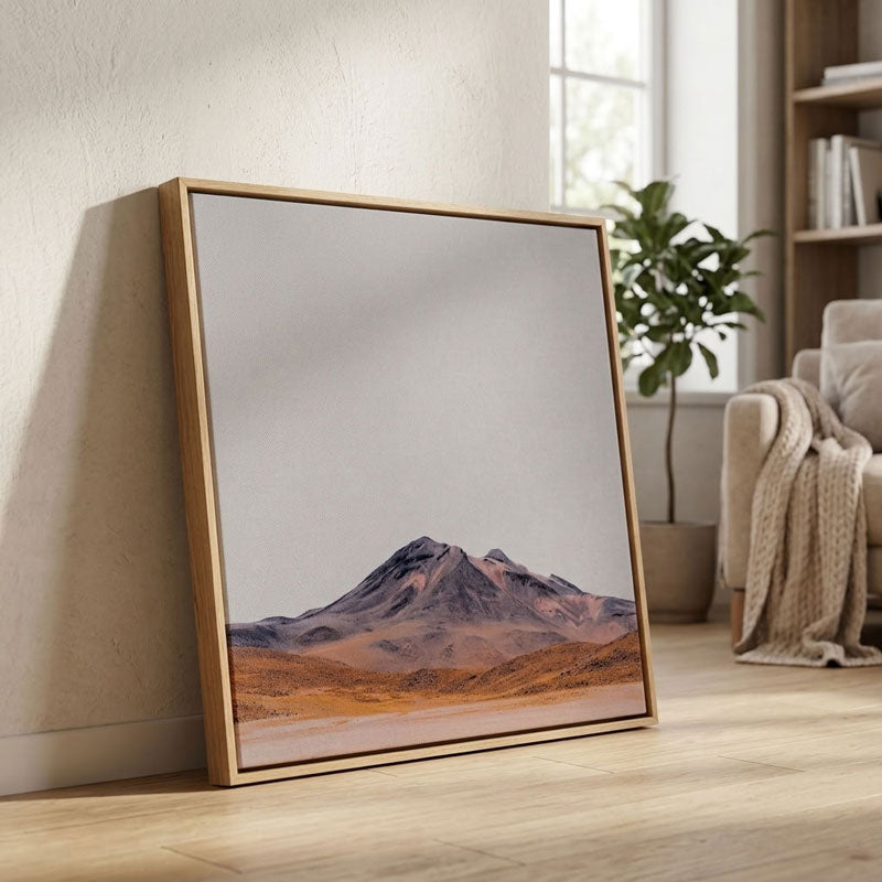 A framed landscape art print leaning against a textured beige wall on a light wood floor, showing a mountain scene with warm brown and orange tones under a pale sky. A sofa, plant, and bookshelf are softly visible in the background.