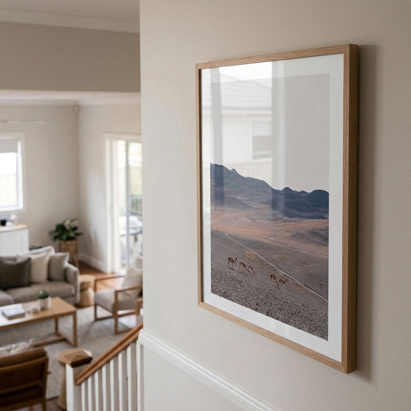 A framed landscape art print featuring a desert scene with mountains and a line of camels, displayed on a beige wall in a modern, sunlit living room.