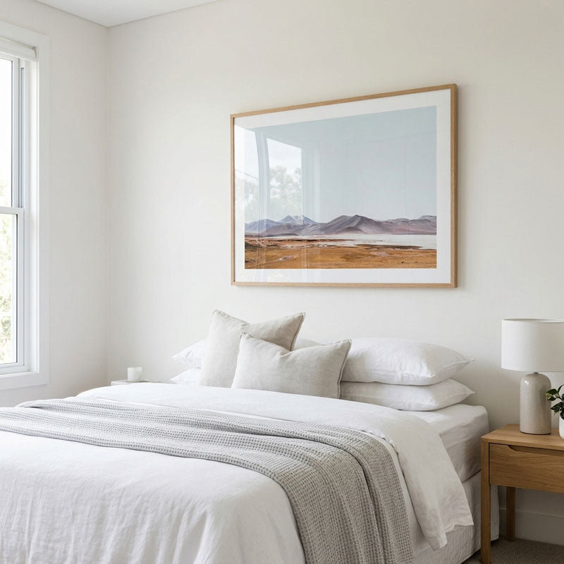 A framed landscape art print hangs on a white wall above a neatly made bed with white linens and a gray textured throw. The room has a light wood nightstand with a white lamp on the right and a window on the left.