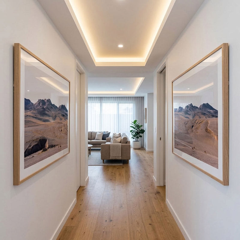 A modern hallway with light wood flooring and white walls, featuring two large framed landscape prints of a desert mountain scene on the left and right walls. Through the hallway, a living room with a beige sofa and a green plant is visible.