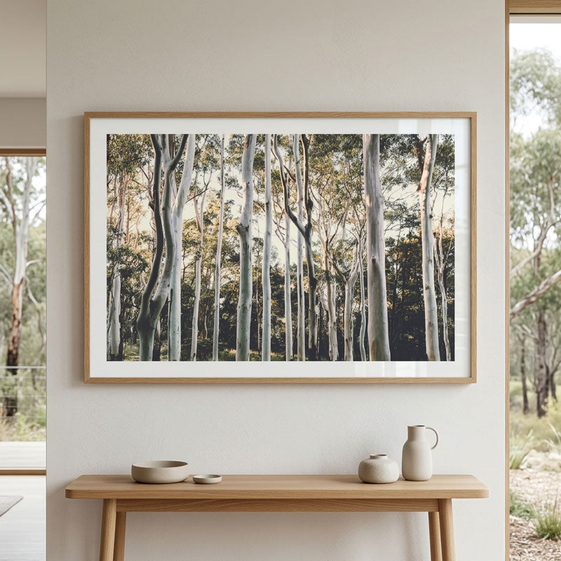 A framed forest art print with tall, slender tree trunks hangs on a light beige wall above a wooden console table with minimalist ceramic decor.