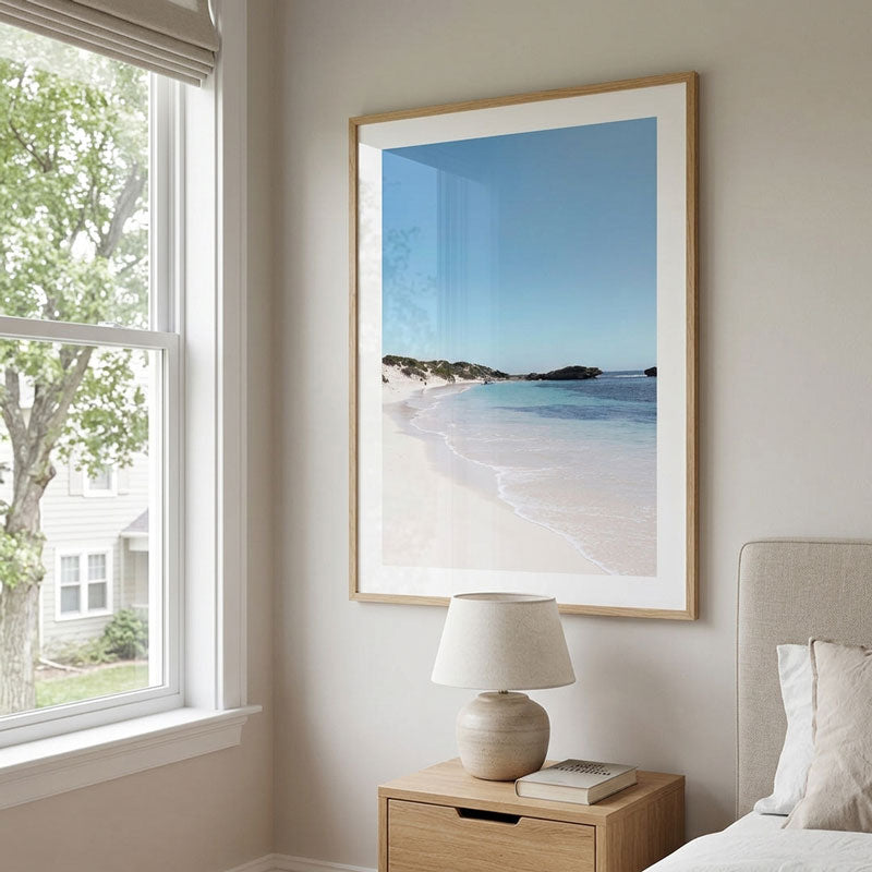 A framed beach photograph with a light wood frame and white mat, displayed on a beige wall above a wooden nightstand with a lamp in a styled bedroom setting.