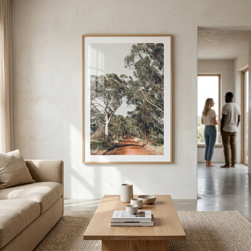 A large framed art print depicting a scenic dirt road lined with tall trees, hanging on a textured off-white wall in a modern, sunlit living room with a beige sofa and a wooden coffee table.