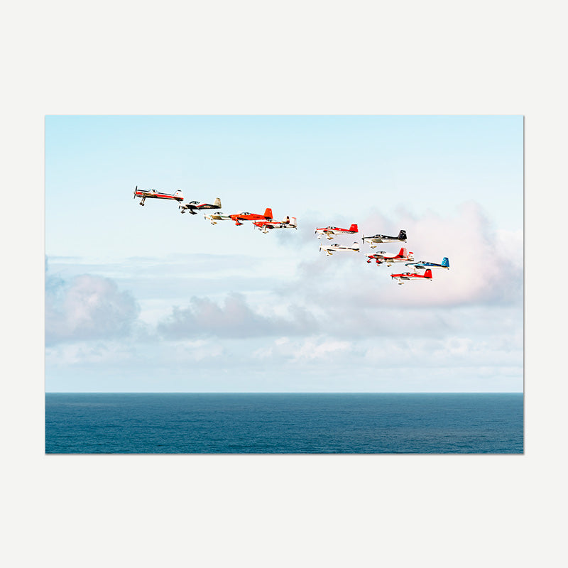 A rectangular wall art print showing a formation of small red, white, and blue airplanes flying over a calm blue ocean under a pale sky with soft clouds.