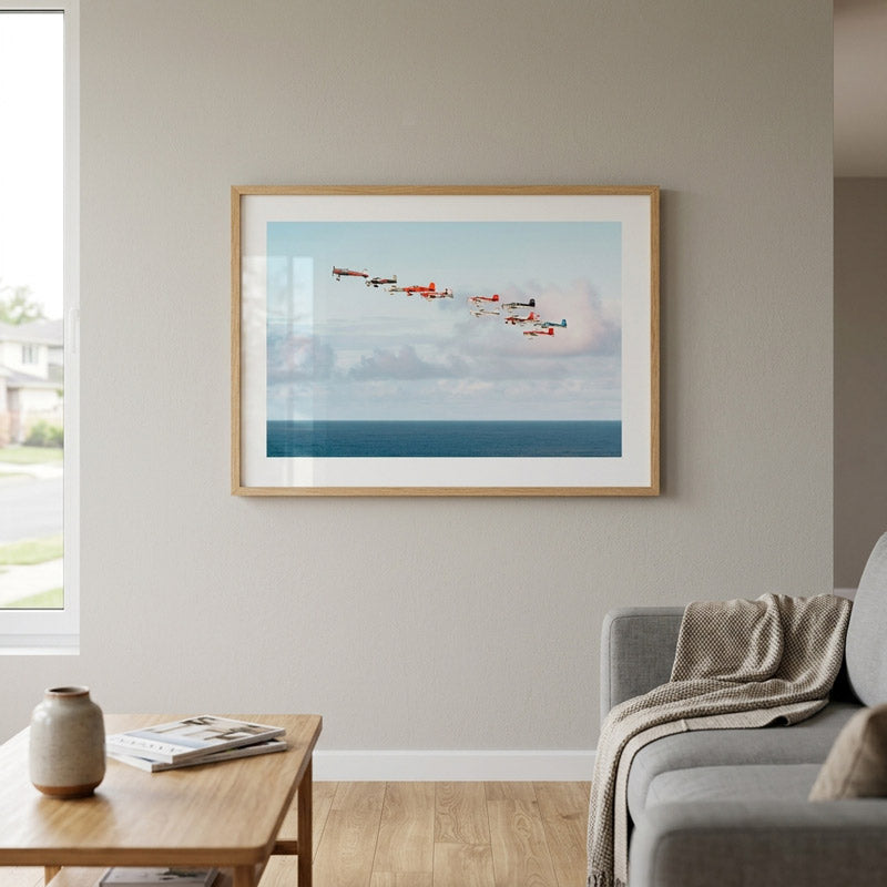 A framed art print depicting a formation of red and white airplanes flying over a blue ocean under a cloudy sky, displayed on a light gray wall in a modern living room setting.